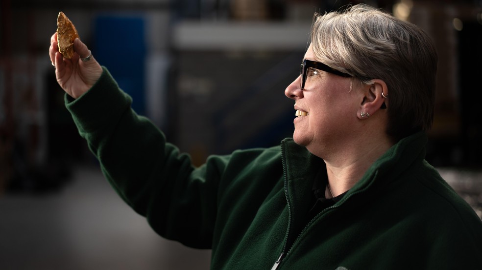 Sara Machin, responsável pelas descobertas da Access +, que trabalha no arquivo de pós-escavação, segurando um machado de mão do período Paleolítico — Foto: BBC News fonte