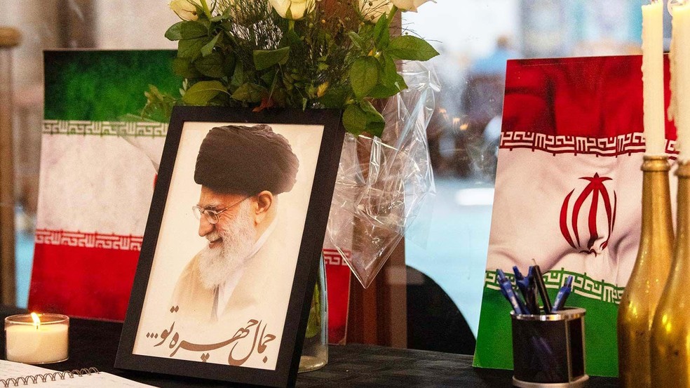 Retrato do líder supremo do Irã, o aiatolá Ali Khamenei, é exibido ao lado de bandeiras iranianas, velas e um livro de condolências em uma mesquita na Cidade do Cabo — Foto: BBC News fonte